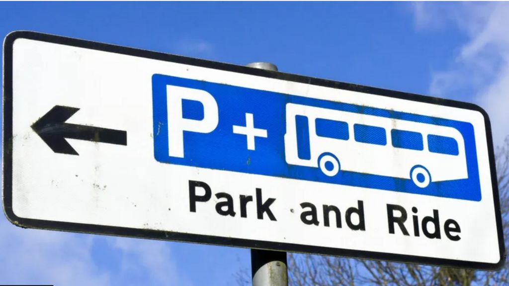 A white rectangular sign with an arrow pointing left and next to it a blue rectangular inside the boundary of the sign with a large letter P (in white), a plus sign (in white) and a bus outlined, in white - and beneath the words "Park and Ride"