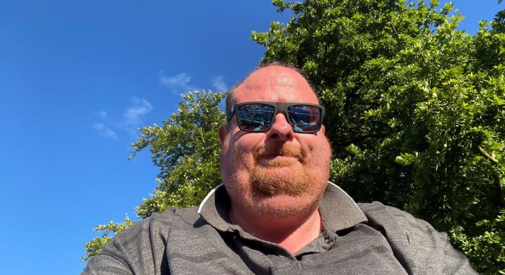 Selfie of Richard Adams wearing black sunglasses with a ginger bread and grey shirt, with a blue sky and tree in the background