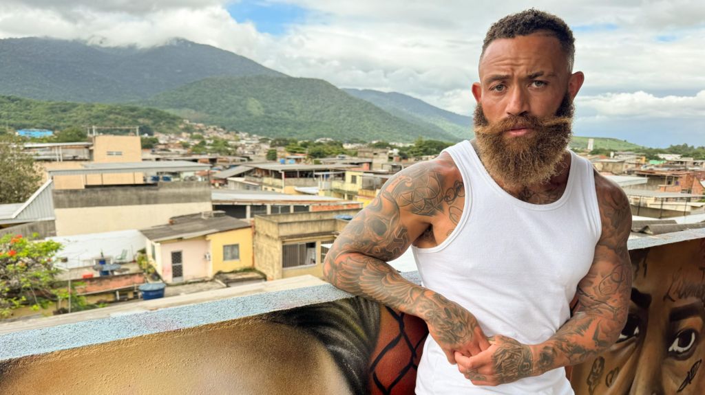 A bearded Ashley Cain looking directly into the camera while filming his BBC documentary series in Rio de Janeiro