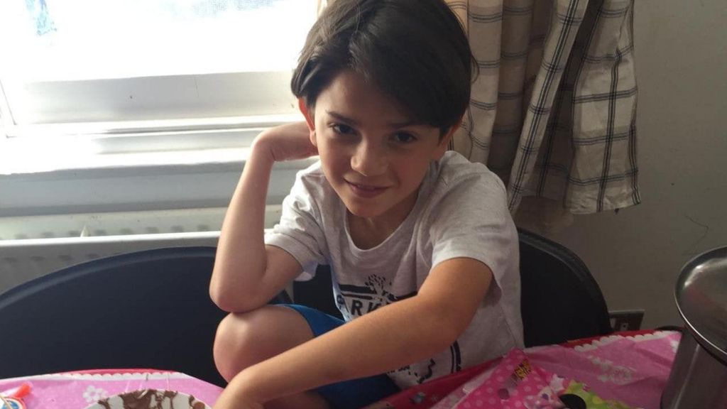 Oliver Archer as a young boy. He is sat on a chair, wearing a grey t-shirt and blue shorts. He has brown hair. 