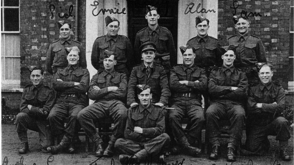 13 soldiers, seated and standing, pose in front of a doorway. The East Dorset Scout Section of Auxiliary Units in 1943, drawn from the Dorset Regiment. Most of the men in this photo joined the SAS and were part of Operation Bulbasket. 