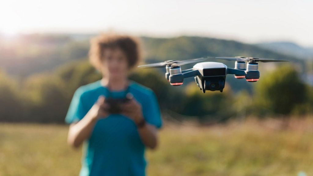 A man flies a drone in a field, surrounded by hills and greenery on a sunny evening.