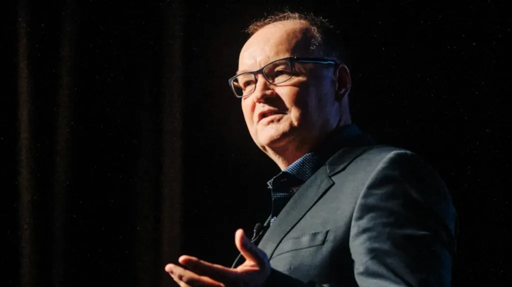 Tony Moretta pictured on stage giving a talk in a darkened room. He is wearing a dark suit and glasses.
