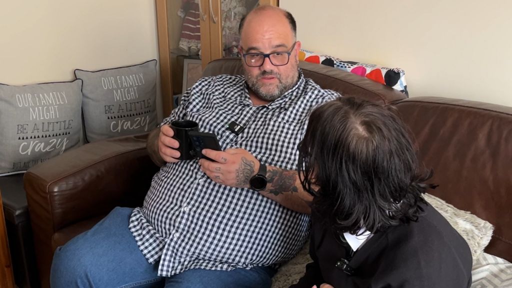 Christopher talking to Lilly on their sofa. Christopher is wearing a blue checked shirt and blue jeans, while holdig a mug and his phone. Lilly has her back to the camera.