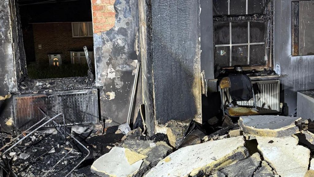 A burnt out room with bits of blackened debris. The wall can be seen split into pieces and scattered across the ground. The windows are also blown out.