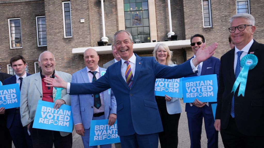 Nigel Farage, a middle-aged man with receding grey hair, wearing a dark blue suit with a coloured stripey tie, stands outside a brown, brick building, with his arms out raised in the air. 