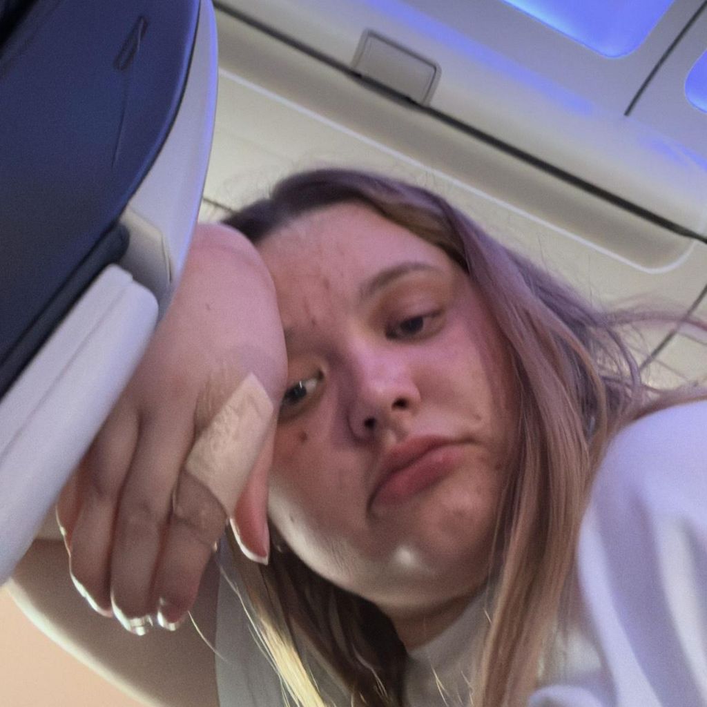 Amy looking exasperated with her head on her hand leaning into the back of the airplane seat after boarding the aircraft