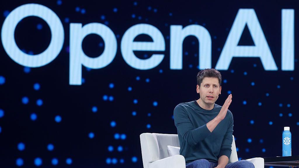 Sam Altman sits in a chair on a stage. He is motioning with his hands. Behind him is the Open AI logo. 