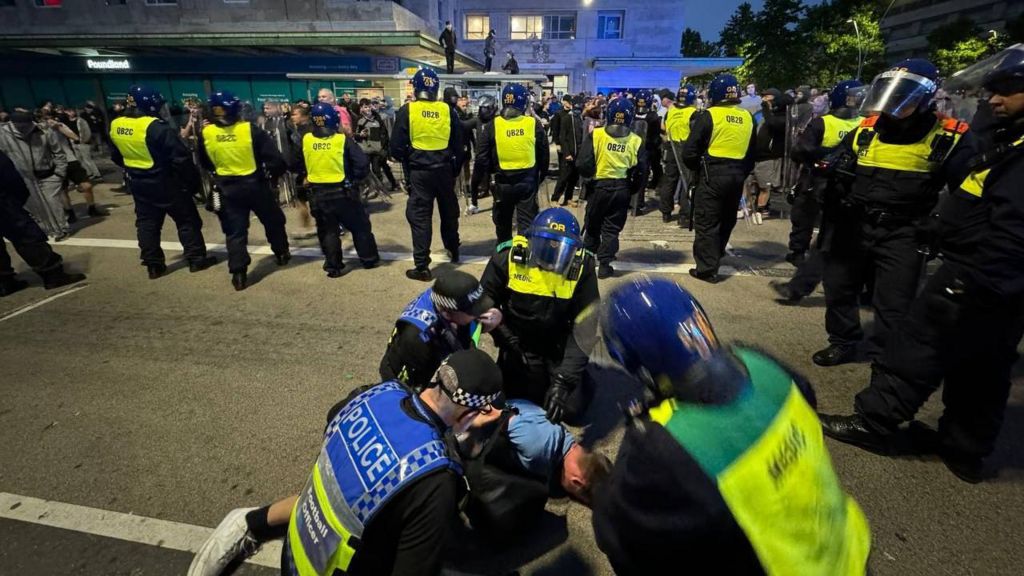 Police officers restrain a man on the floor while other officers control crowds of protesters