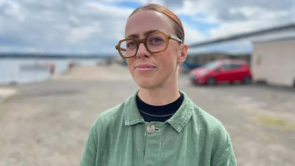 A woman with glasses and blonde hair pulled back wears a green jacket over a dark top, standing outdoors. Behind her is a blurred background showing a bridge structure, cloudy sky, and a red vehicle. The setting appears to be near the Forth Road Bridge.