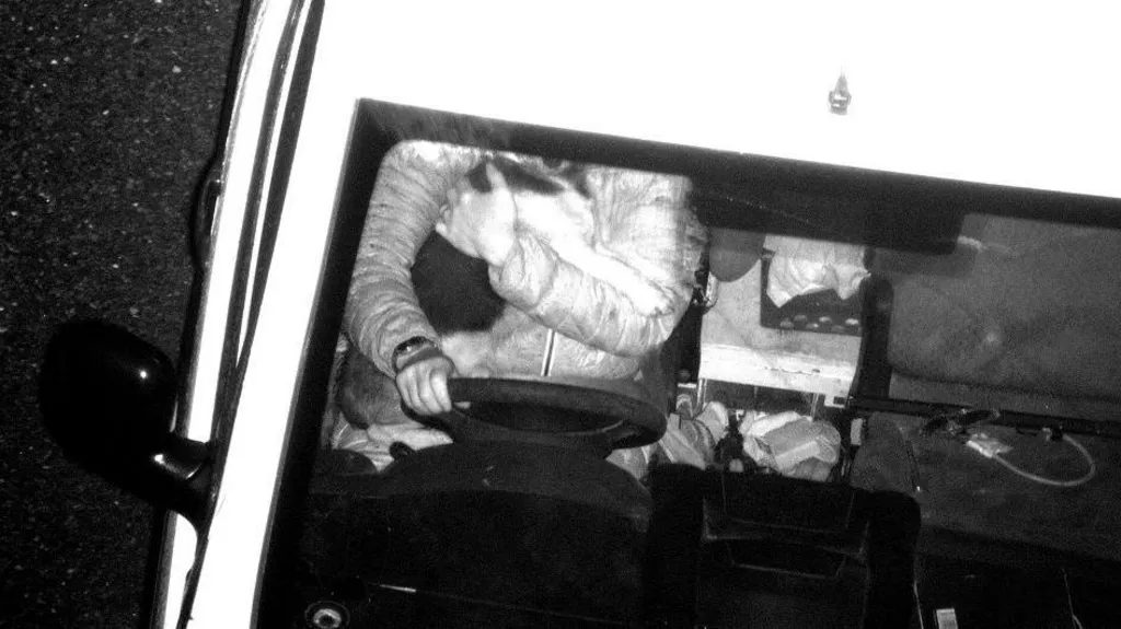 The image shows an overhead, black‑and‑white view of a person driving a vehicle while holding a small dog close to their chest. The driver’s left hand is on the steering wheel and their right arm is wrapped around the dog. The dog’s face is pressed up near the driver’s face.