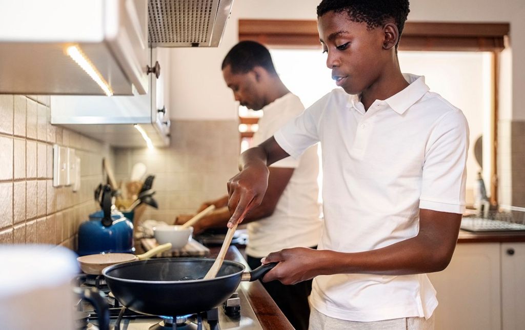 Stock photo showing a teenager using a fying pan while cooking in the kitchen with his father in the background