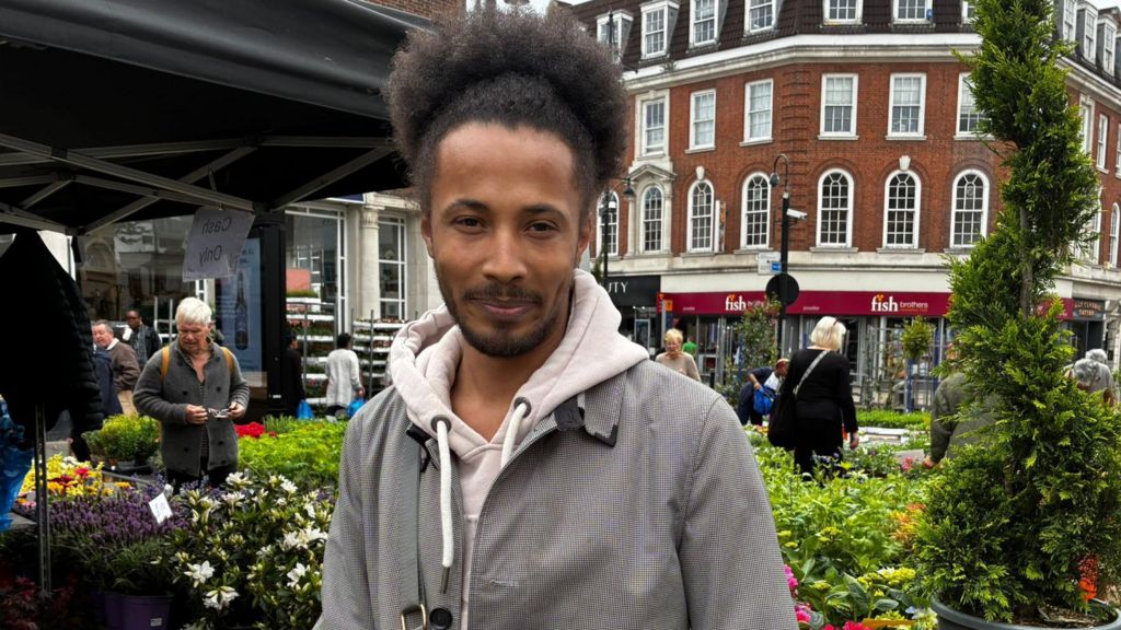 Josh is seen standing in front of a flower stall. He is wearing a grey jacket over a light-coloured hoodie.