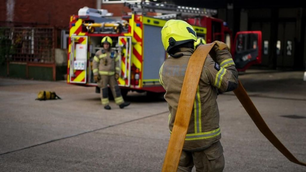 Firefighter officers running through a practice drill during a London Fire Brigade facility. One faces away from the camera holding a hose and another stands near a fire engine in the distance.