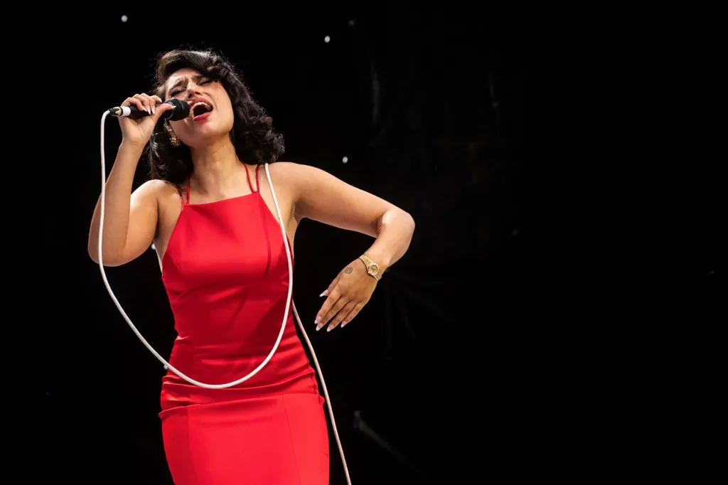 Raye sings on stage wearing a red dress with a microphone in her hand, with the lead over her shoulder 