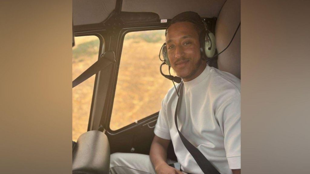 A man in a white shirt sits in a helicopter with a headset on. He smiles at the camera.