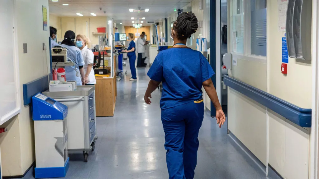 A female nurse is seen from behind walking down a hospital corridor, wearing royal blue scrubs. There are various items of hospital equipment and staff wearing masks further down the corridor.