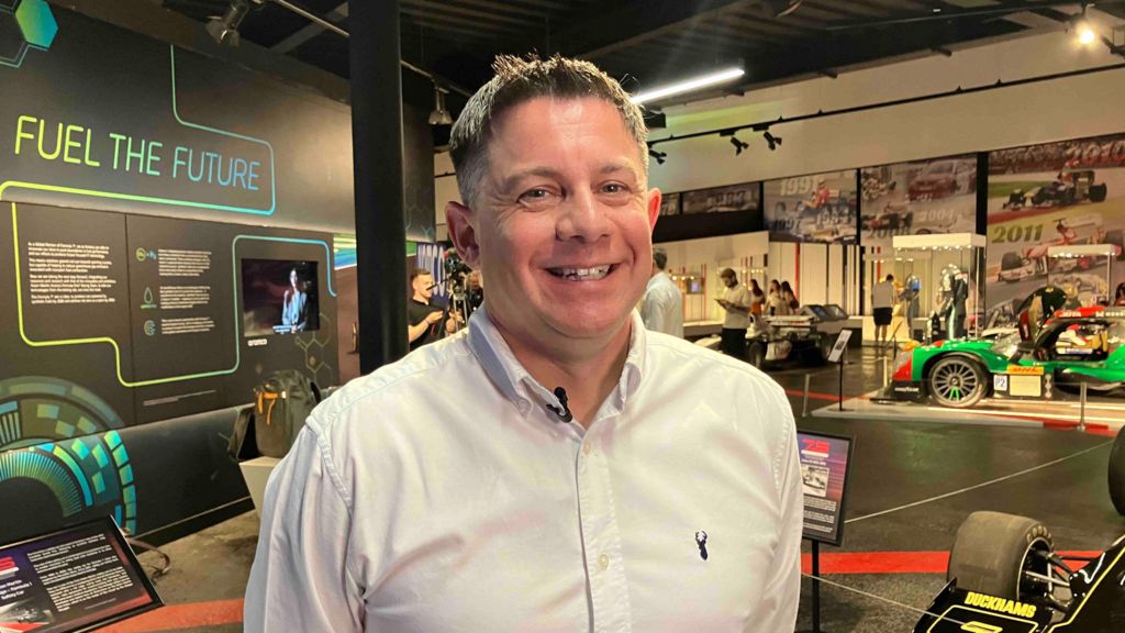 A man in a white shirt stands in a room full of racing car displays.
