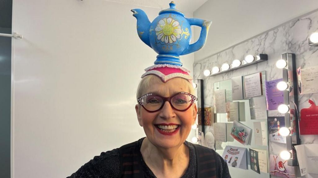 Woman with glasses smiles at the camera. She has a blue teapot on her head and a mirror with Christmas card on in the background.