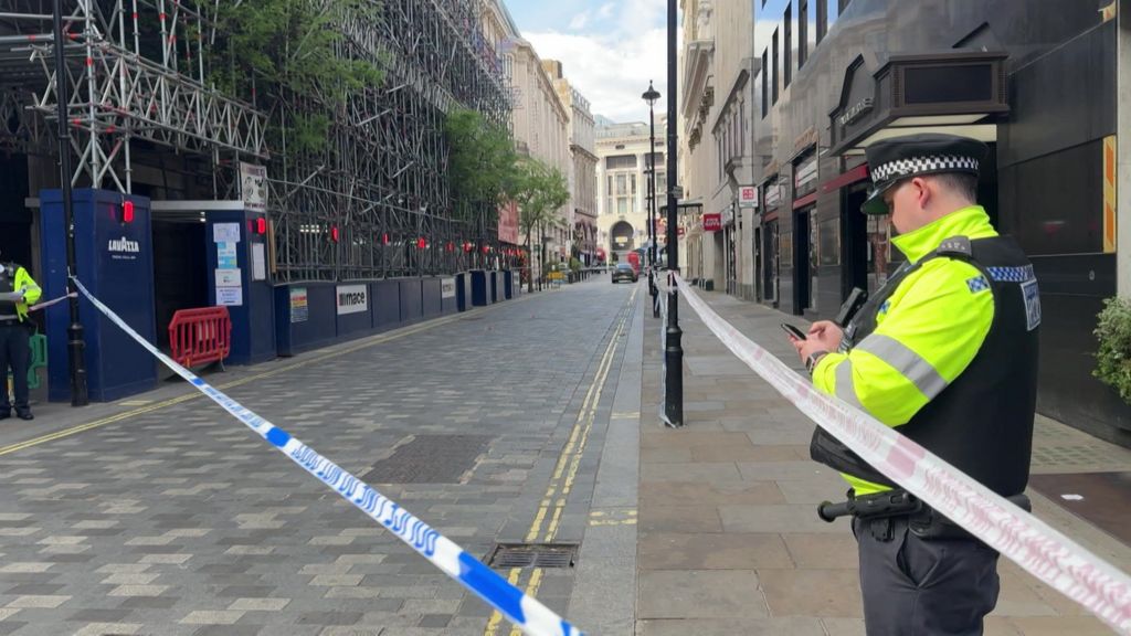 Police tape blocks off a central London street as officers stand on either side of the cordon near scaffolding and shopfronts, with the largely empty road stretching into the distance.