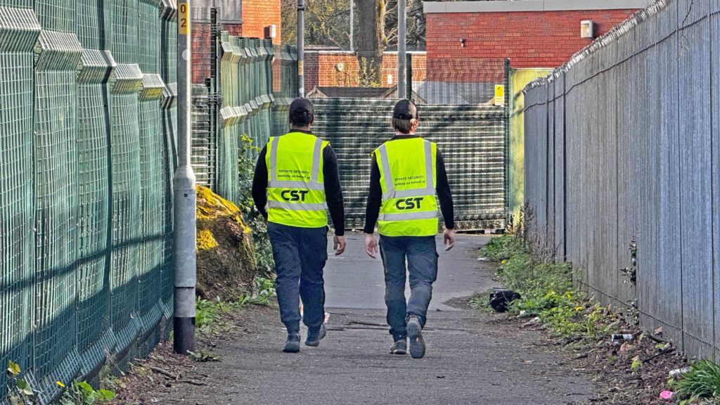 Two men wearing black caps, high-viz waistcoats and dark trousers walk away from the camera down a wide footpath, with high fences either side.