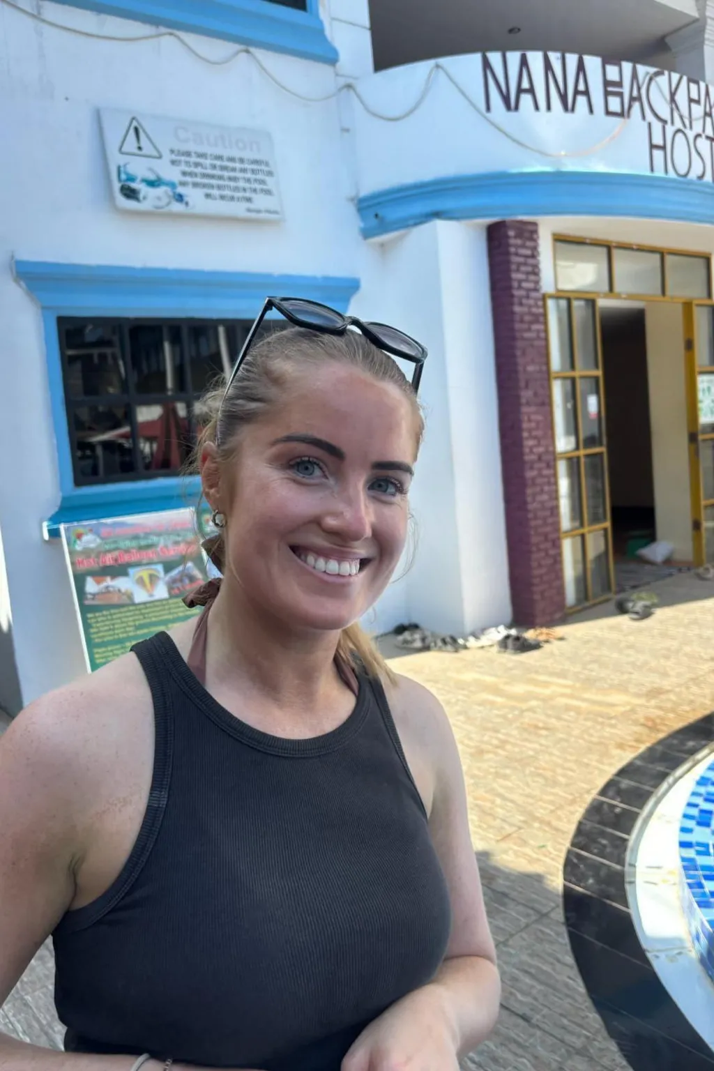 A young woman in a black vest top, hair tied back and sunglasses resting on her head, stands outside a white building with a crude 