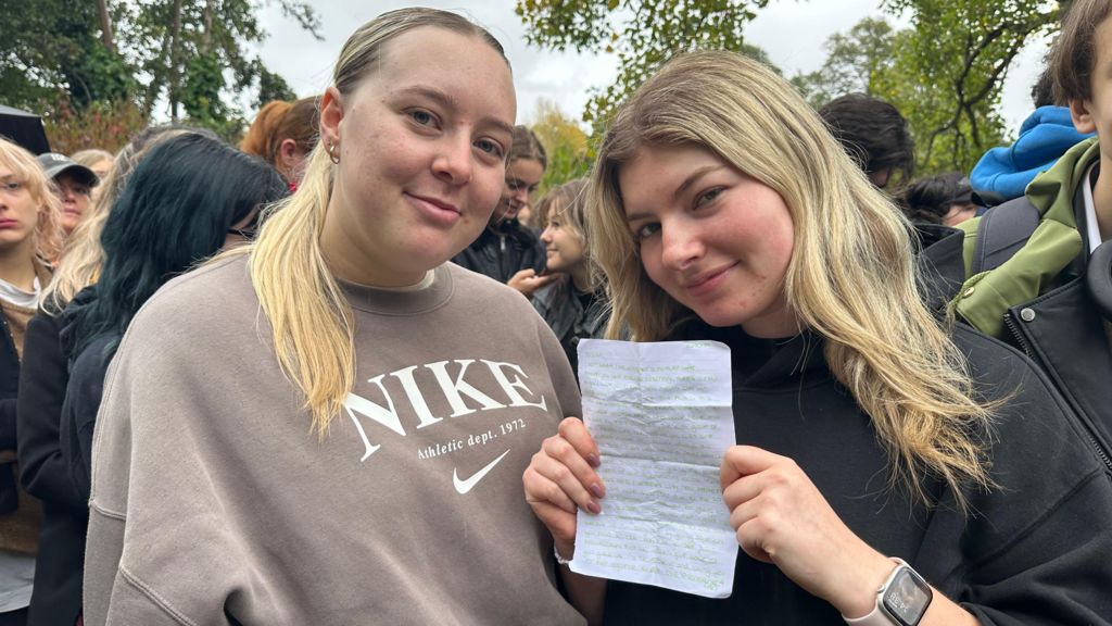 Emily and Olivia pose for the camera while stood in a crowd at Hyde Park. One of them is holding a handwritten note. They both have blonde hair and are smiling.