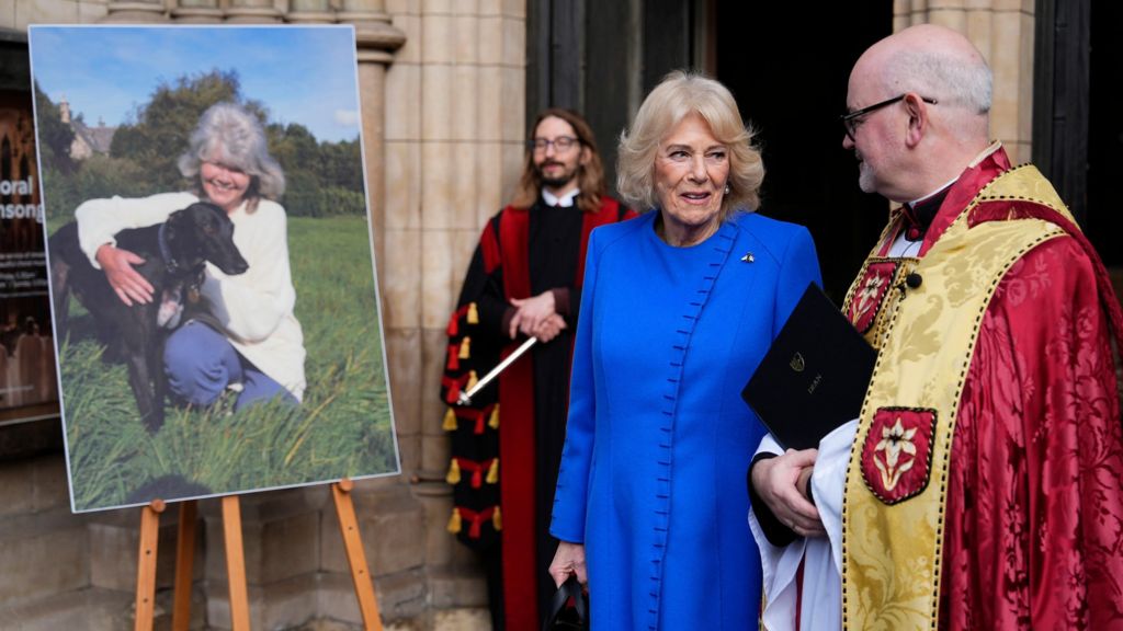 The Queen speaks to the Dean while both stand next to a printed image of Jilly Cooper