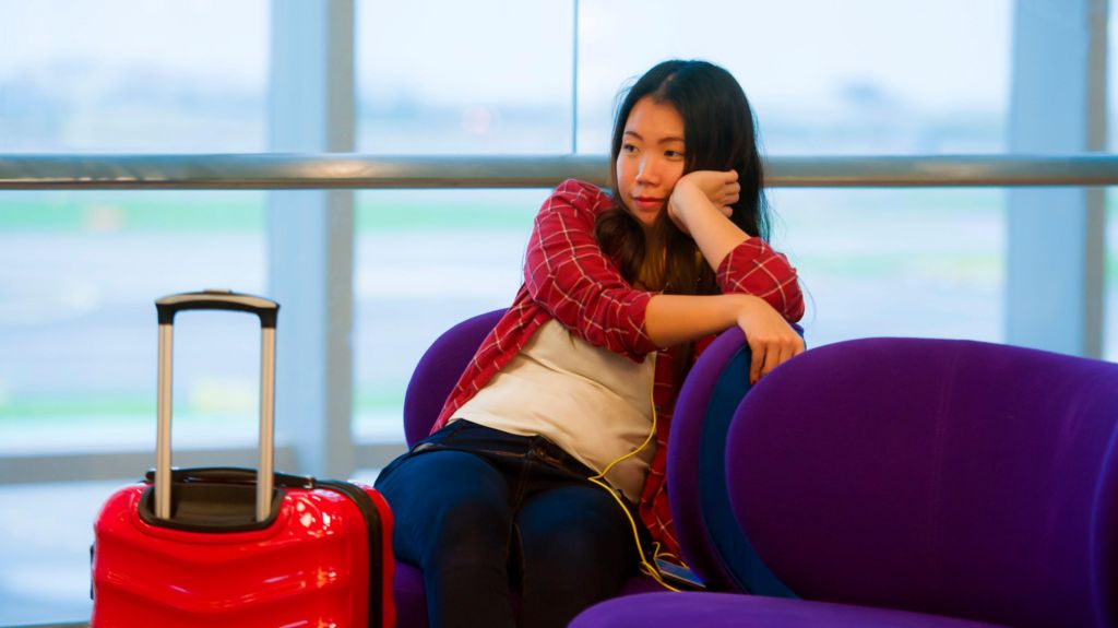 Young woman in red checked shirt rests her chin on her hand. She is sitting in an airport waiting room with a red case next to her