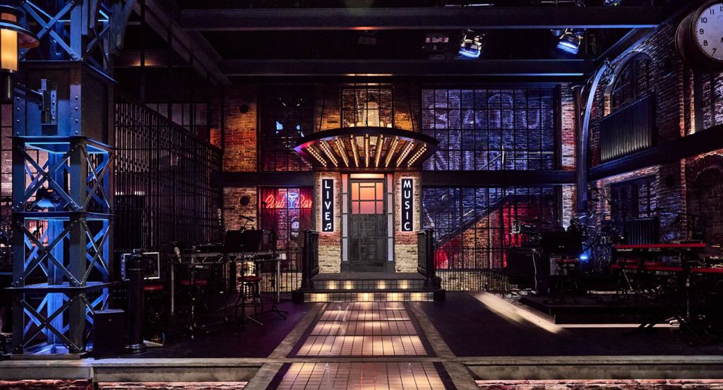 The SNL UK set as seen from the view of the audience and cameras, with live band instruments set up either side of a central walkway and a brickwork-style surrounding background, illuminated in dark shades of blue, orange and red.