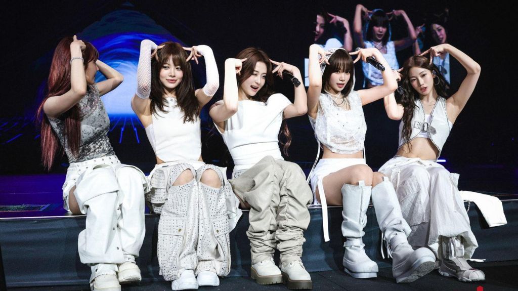 Le Sserafim sit on stage during their EasyCrazyHot tour, making 'cat ears' with their fingers, while wearing toned grey and white street wear