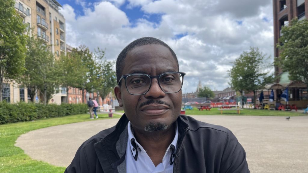 Israel Eguaogie, wearing black glasses and a black jacket, sits in a public square in Belfast