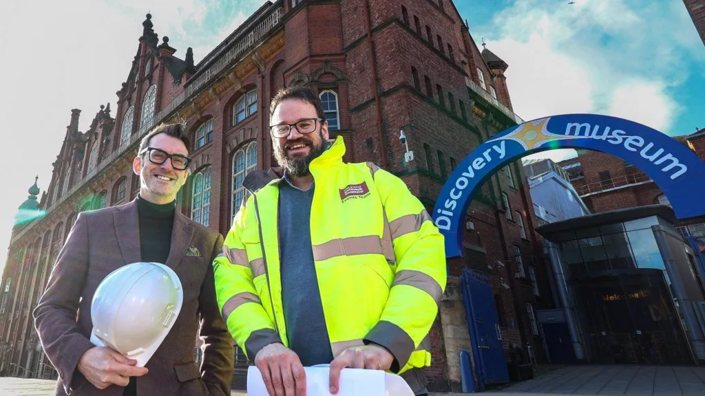 Keith Merrin, Director of North East Museums and Nick Butterley, Customer & Facilities Manager of Discovery Museum are smiling outside the museum while holding protective white helmets. Merrin is wearing a brown tweed jacket and trousers with a black turtle neck and glasses. He has short brown hair. Butterley is wearing a high visibility jacket on top of a blue jumper. He has glasses, short brown hair and beard. The blue arched sign reading Discovery Museum at the venue's entrance is next to him.