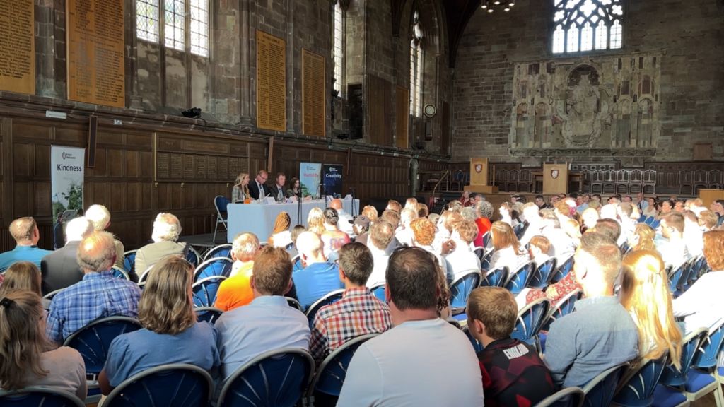 A crowd of people seated at an election hustings in a hall, with the panel at the front of the room 