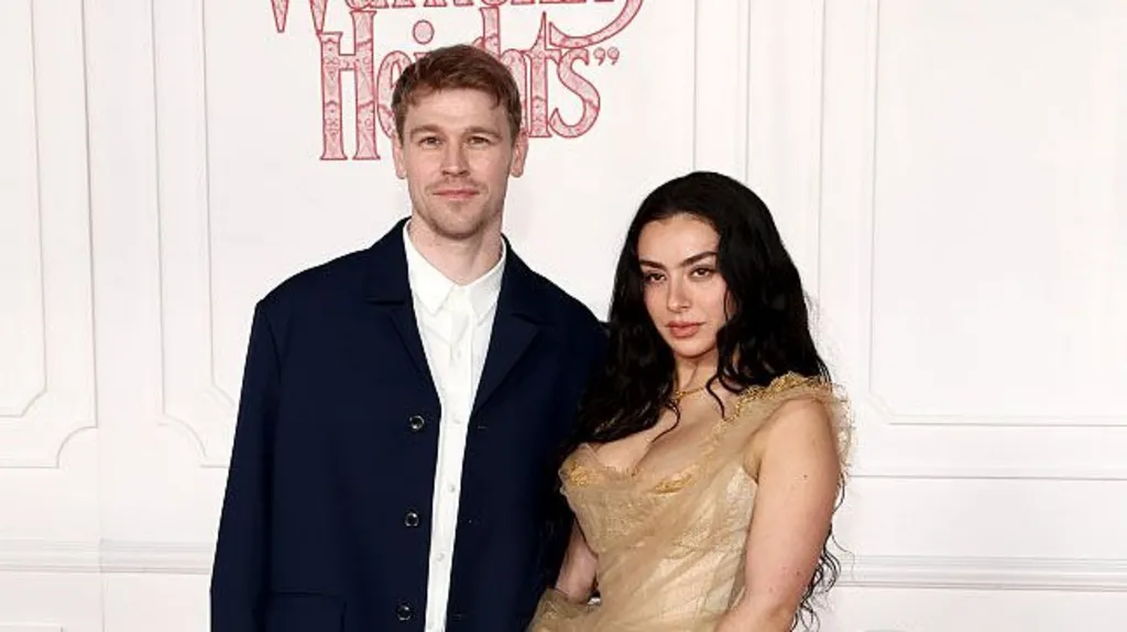 A close-up photo of Finn Keane and Charli XCX stood next to each other on a red carpet. There is a white wall behind them and Finn's head is blocking the word Heights from a Wuthering Heights logo. Finn is wearing a white shirt with a dark navy jacket unbuttoned and is staring directly at the camera. Charli has her long black hair down and is wearing a gold dress.