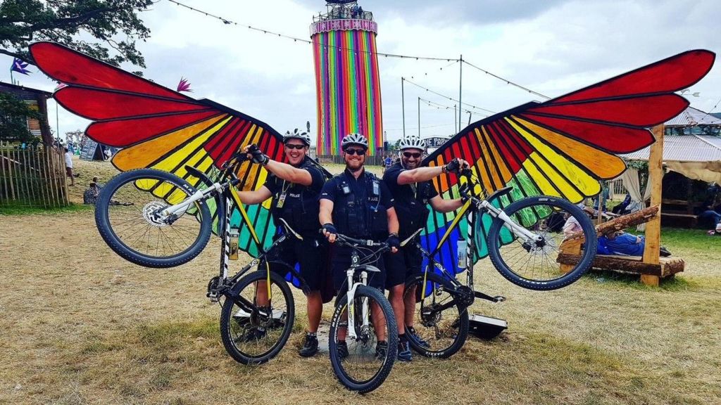 Three policeman and their bikes posing in front of the large colourful wings at Glastonbury