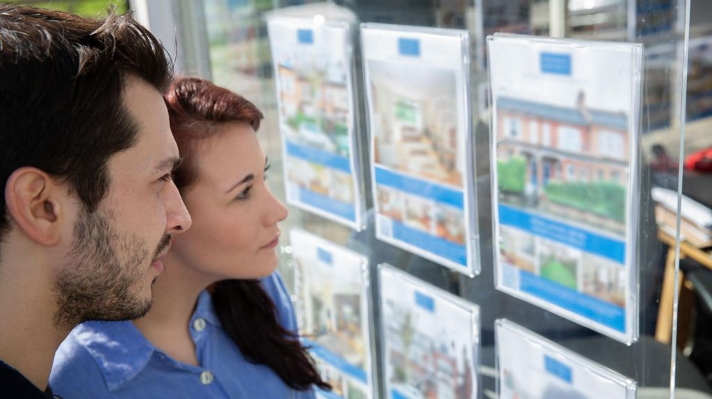 A young man and woman stand alongside each other looking at an estate agents' window.