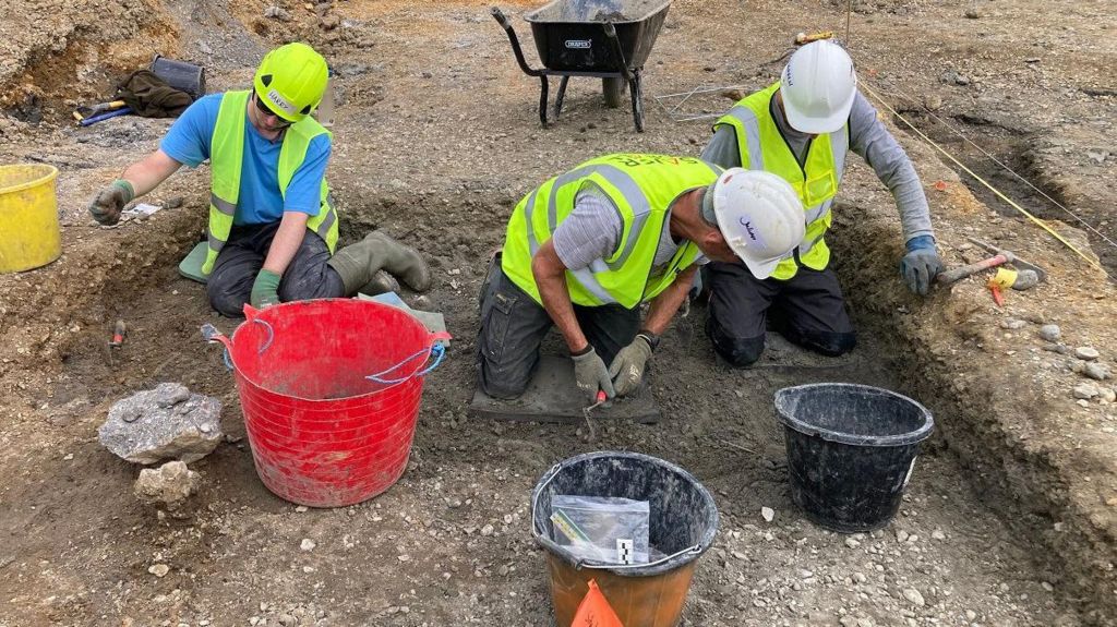 Three people wearing green high vis vests and hard hats kneeling on the ground