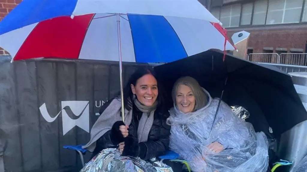 Michelle Power and Karen Dunkley, two women, one in a black coat and grey scarf is grinning while holding a blue, red and white umbrella and the other woman in wearing a translucent poncho and holding a black umbrella