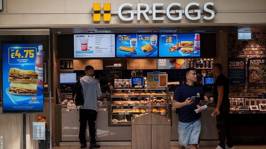 A customer exits a Greggs bakery with a meal deal while another shopper pays at the bakery chain's till