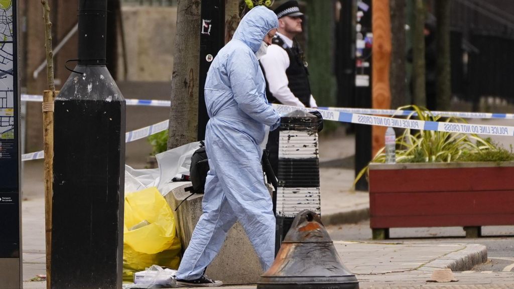 A forensic investigator at the scene on Chalton Street, Camden, walking along a police cordon on the pavement. A police officer can be seen standing in the background.