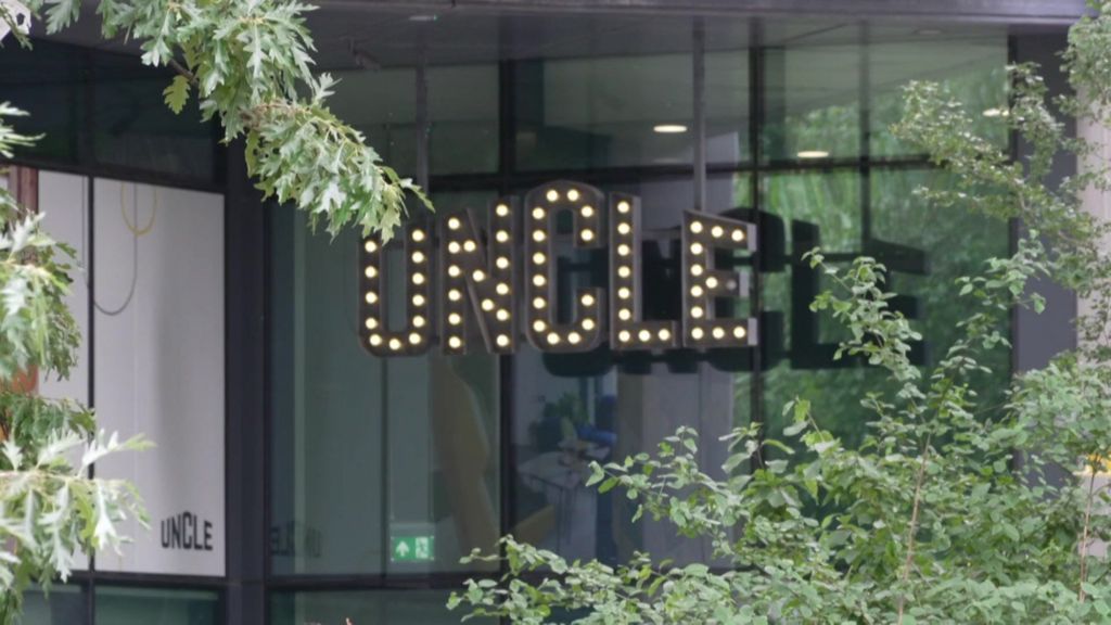 Building entrance featuring a prominent, illuminated marquee-style sign that spells out the word "uncle" in glowing light bulbs, partially framed by green foliage.