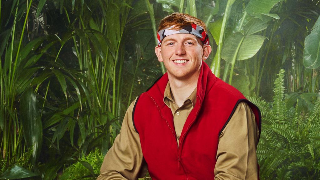 AngryGinge wears a red vest and tan shirt and a headband in front of a wall with forest leaves on it.