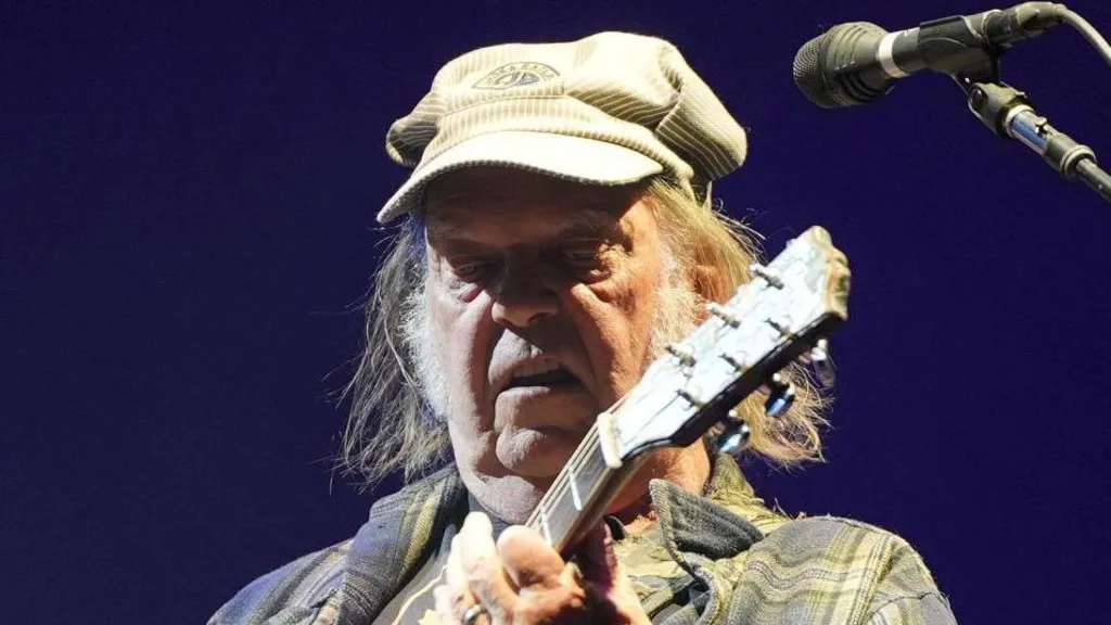 Neil Young is playing a guitar on stage while wearing a green cap and checkered jacket. He has long hair and is standing next to a microphone.