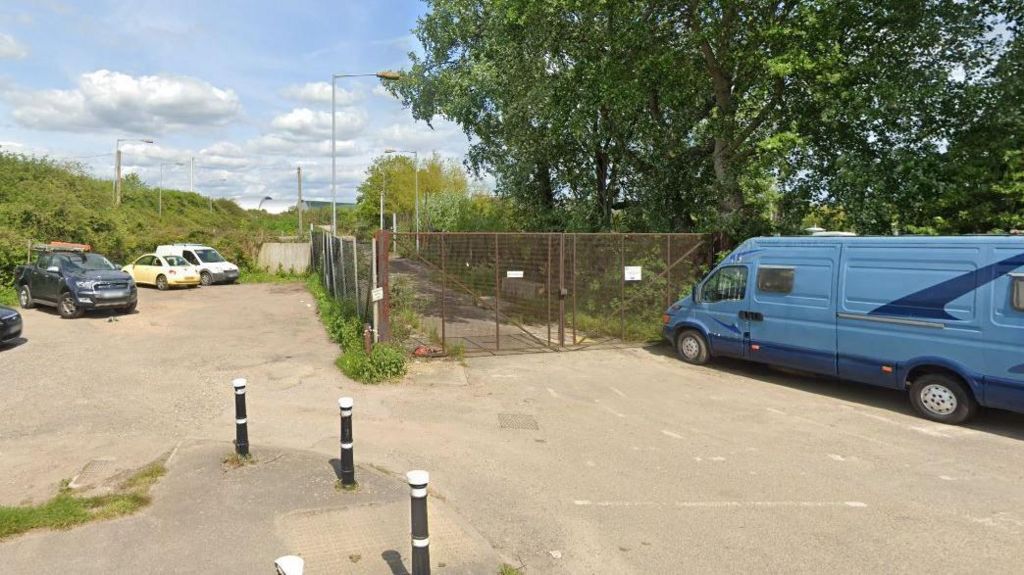A google maps image of the rusted gates of a former recycling centre, with bollards and a dirt carpark outside and a blue van parked at the gates