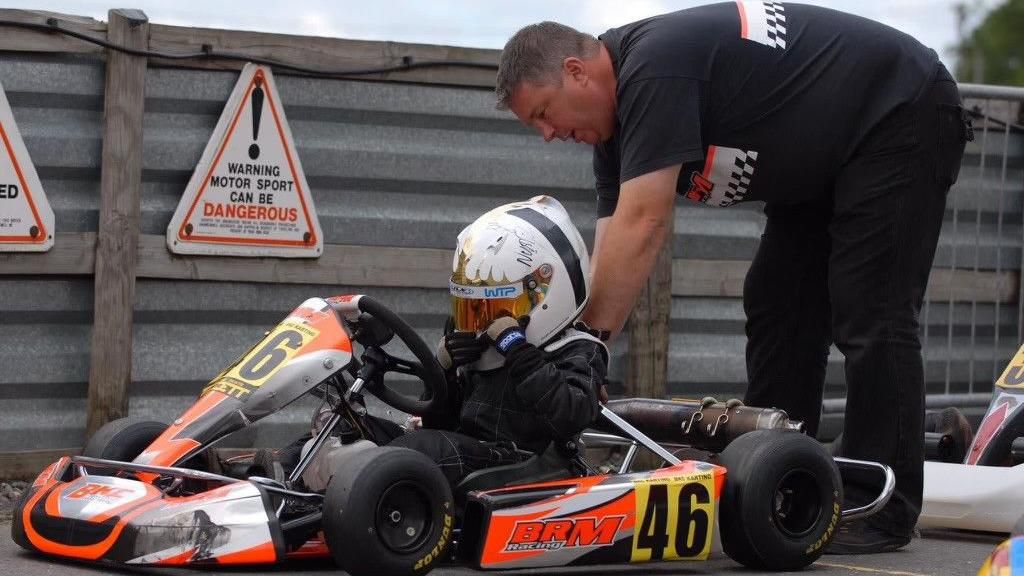 Image of Rob Dodds with Lando Norris as a child. Rob is helping Lando, who is sat behind the wheel of a bright orange race car. Lando is wearing a white helmet