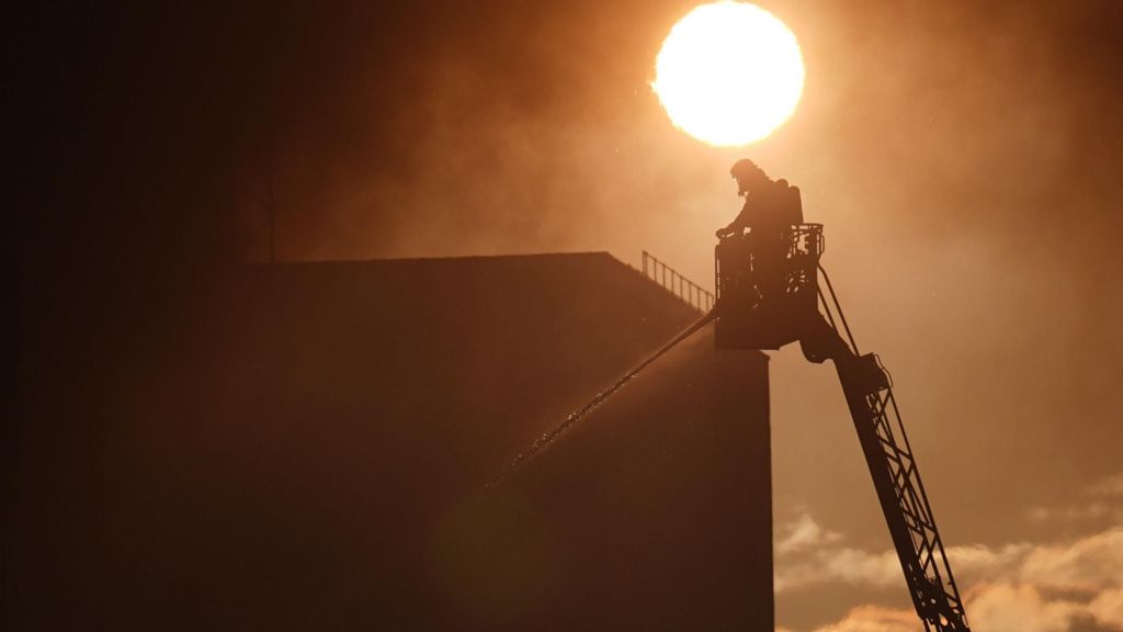 Silhouette of a firefighter in a raised mechanical crane platform spraying a stream of water downwards against a thick blanket of smoke. The bright sun glows through the hazy, orange sky above the dark silhouette of a building.