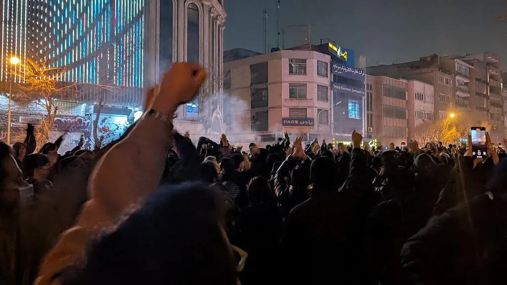 Hundreds of people stand in the streets of Tehran with their fists raised in the air. There is a small amount of smoke hovering above their heads in parts of the picture.