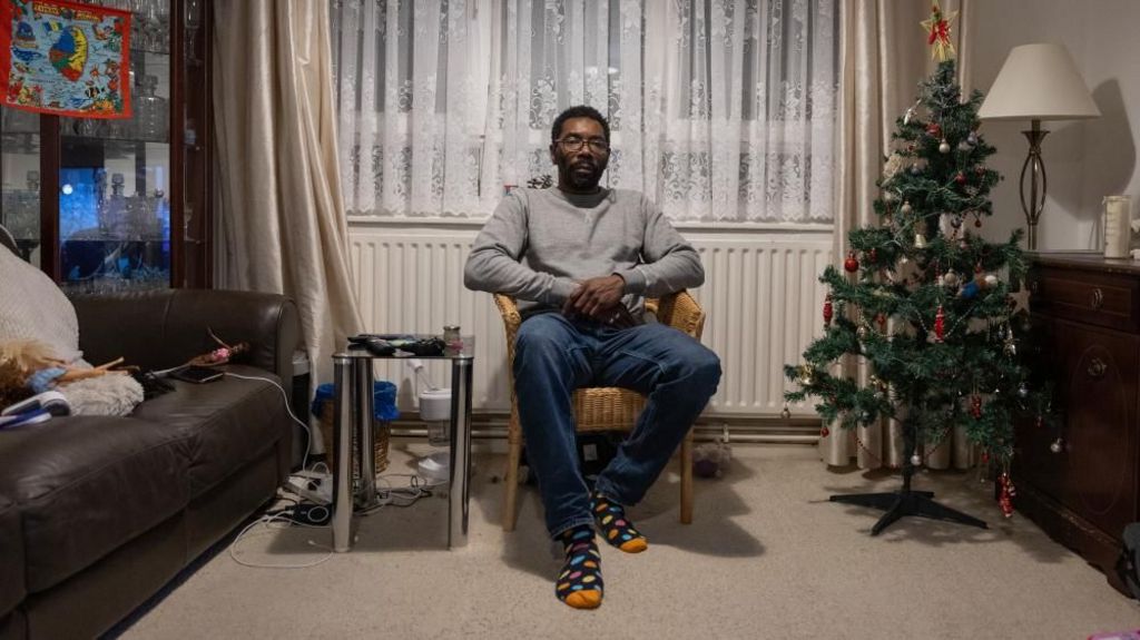 Man sits in a rattan chair in the living room with a decorated Christmas tree to the right hand side of him. To the left is a brown sofa and a small table.