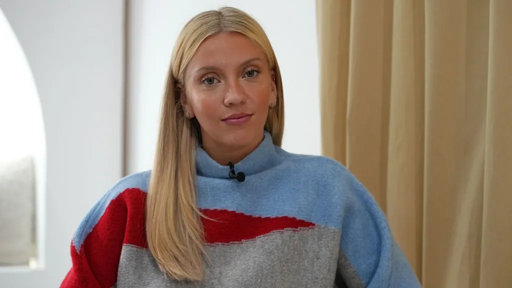 A young woman sits near a window and in front of a curtain. She is wearing a blue, red and grey knitted jumper and has long blonde hair - she has small hoop earrings in both ears. Her face is tanned.